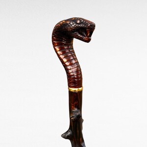 May include: A handcrafted walking cane featuring a cobra head handle. The handle is carved and painted in brown and black, with a gold band. The shaft is dark brown, textured wood. A unique and decorative item.