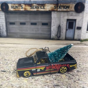 May include: Black pickup truck with a small artificial Christmas tree in the bed. The truck has "Hot Wheels" written on the side and is a Christmas ornament.