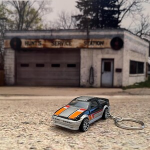 May include: A black and silver toy car with orange and white racing stripes. The car is a keychain and is sitting on a gray surface in front of an old, abandoned service station with the words "Hunt's Service Station" on the front.