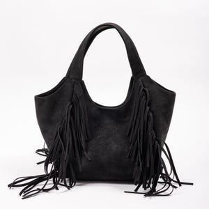 May include: A black suede handbag featuring long fringe detailing on the front panels and two sturdy shoulder straps. The word baggu is embossed in the centre of the bag. The design includes a curved top opening and a structured base.