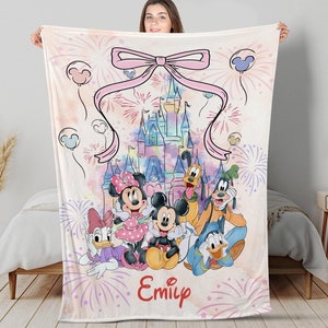 Puede incluir: Una manta suave y clara con un diseño de acuarela de personajes de Disney, incluyendo Mickey y Minnie Mouse, y un castillo. La manta tiene un lazo rosa y el nombre "Emily" en escritura roja.