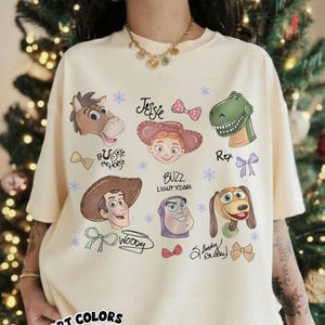 May include: Ivory-colored t-shirt featuring cartoon characters from the Toy Story franchise. The design includes images of Woody, Buzz Lightyear, Jessie, Rex, and others, with their names and decorative bows. The shirt also has the words "COMFORT COLORS" and "Ivory".