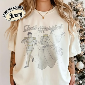 May include: An ivory t-shirt featuring a graphic illustration of a prince and a princess dancing. The text "Just Married" is printed above the figures in a cursive font. The design includes small star accents surrounding the couple.