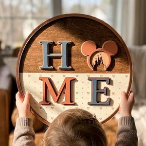 Könnte beinhalten: Ein rundes Wand-Schild aus Holz mit dem Wort HOME in erhabenen, mehrfarbigen Buchstaben. Der Buchstabe O ist durch eine Mausohr-Silhouette mit einer Schloss-Ausstanzung ersetzt. Die obere Hälfte ist aus dunklem Holz, die untere ist cremefarben.