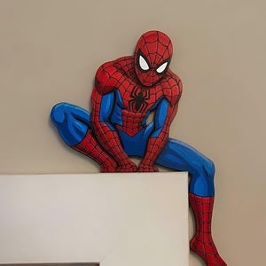 May include: A flat, cutout wall decoration of the superhero Spider-Man in a crouching pose. The figure features the iconic red and blue suit with a black web pattern, a black spider emblem on the chest, and large white eyes with black outlines.