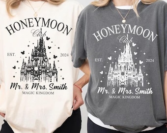 May include: Two t-shirts, one ivory and one pepper-colored, feature a graphic design for the "Honeymoon Club." The design includes a castle illustration, the text "Mr. & Mrs. Smith," and the year "2024."