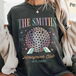 Op de afbeelding: Donkergrijs t-shirt met de tekst "THE SMITHS" boven een kleurrijk geometrisch bolontwerp. Onder de bol staat de tekst "Honeymoon Club EPCOT, FLORIDA" met het jaar "2024" en Mickey Mouse-iconen.