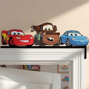 May include: A black L-shaped door topper featuring three cartoon car characters: a red race car with the number 95, a brown rusty tow truck, and a light blue sports car. Each character has large expressive eyes and a smiling mouth.