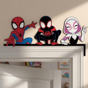 May include: A black door frame topper featuring three cartoon characters: a red and blue Spider-Man, a black and red Miles Morales Spider-Man, and a white, pink, and black Spider-Gwen. The characters are positioned in a row along the top edge.
