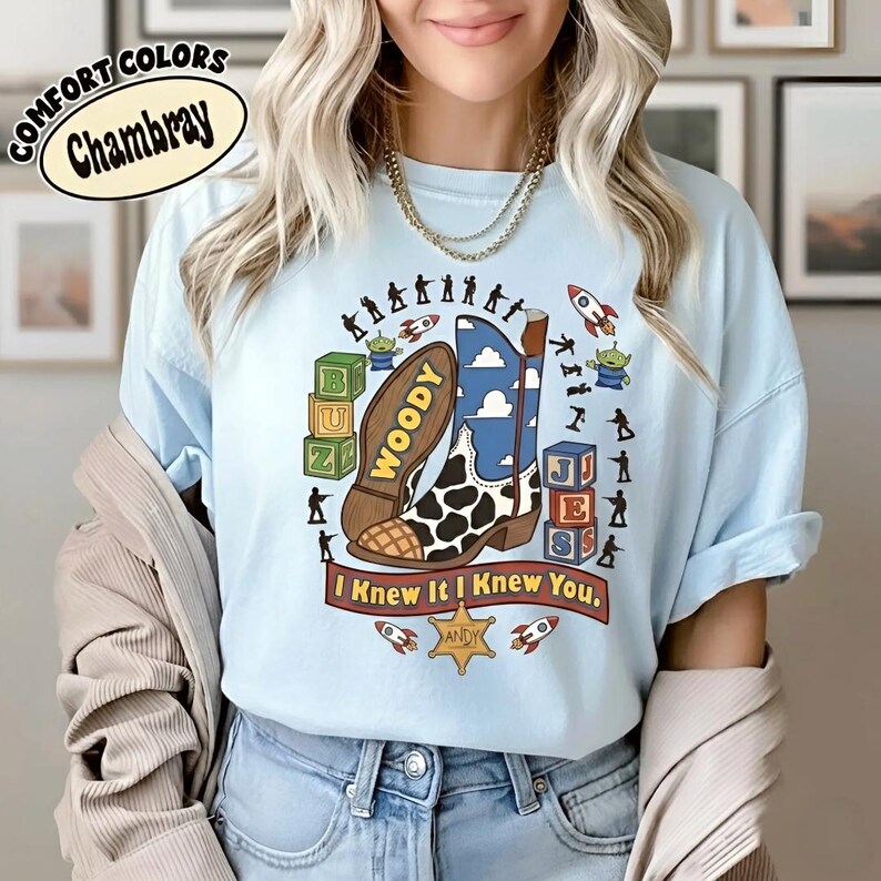 May include: A light blue t-shirt featuring a graphic print of a cowboy boot with a cow-print toe and blue sky pattern. The design includes toy blocks, green aliens, rockets, plastic soldiers, a sheriff badge labeled Andy, and the text I Knew It I Knew You.