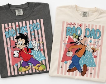 May include: Two crewneck t-shirts in charcoal gray and cream. The charcoal shirt features Max Goof with the text RAD LIKE DAD. The cream shirt features Goofy with the text RAD DAD. Both shirts display 90s-style pink stripes, geometric shapes, and blue accents.