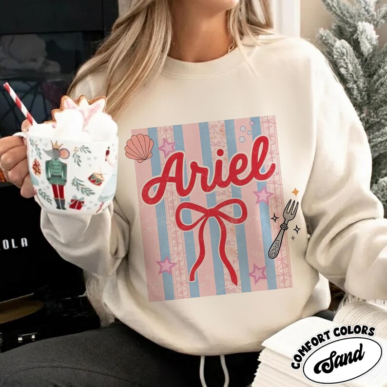 May include: Cream-colored sweatshirt with a graphic featuring the name "Ariel" in red script, a red bow, and a fork illustration. The design includes pink and blue stripes, stars, and a seashell. The sweatshirt has the Comfort Colors brand label.