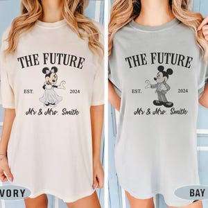 Puede incluir: Dos camisetas, una de color marfil y la otra azul claro, con un gr&aacute;fico de Mickey y Minnie Mouse vestidos con atuendos de boda. El texto en las camisetas dice "The Future Est. 2024 Mr & Mrs. Smith".