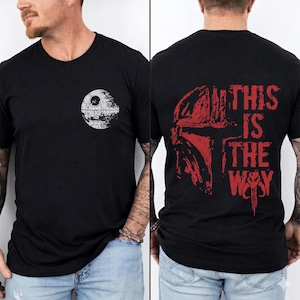 May include: Black t-shirt featuring a white Death Star graphic on the front and a red Mandalorian helmet design with the text "THIS IS THE WAY" on the back. The shirt is a crew neck style.