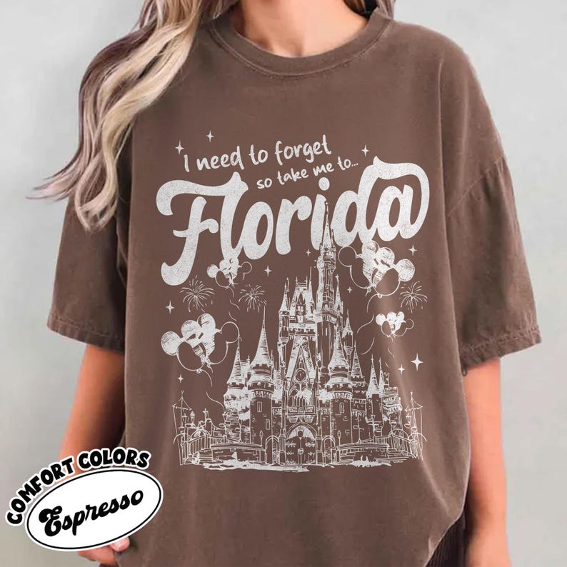 May include: Espresso-coloured t-shirt featuring the text "I need to forget so take me to... Florida" in white script. The design includes a castle illustration, fireworks, and Mickey Mouse balloons. The shirt is from Comfort Colors.