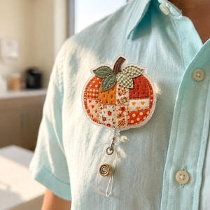 Puede incluir: Un portatarjetas con forma de calabaza que presenta un diseño de patchwork con varios patrones florales y de corazones en naranja, rojo y blanco. La calabaza tiene hojas de cuadros verdes y un tallo marrón, unido a un clip de plástico.