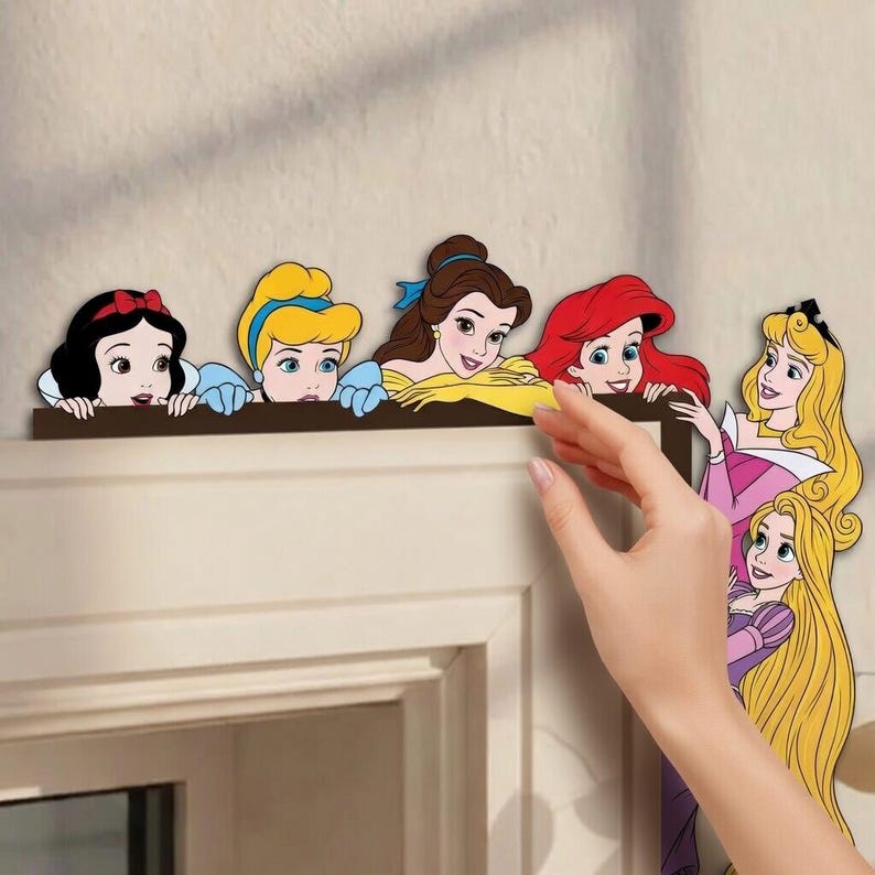 May include: A decorative wall decal set featuring six cartoon princesses. Snow White, Cinderella, Belle, Ariel, Aurora, and Rapunzel are depicted in colorful outfits. The characters are arranged to appear as if they are peeking over a door frame.