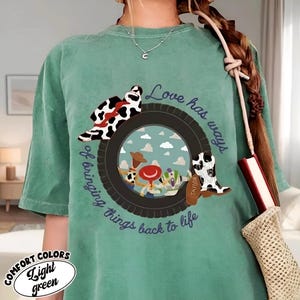 Puede incluir: Una camiseta verde claro con un gr&aacute;fico de un neum&aacute;tico negro que enmarca una escena de cielo con tres personajes de dibujos animados. Un sombrero y una bota con estampado de vaca est&aacute;n sobre el neum&aacute;tico. El texto dice, Love has ways of bringing things back to life.