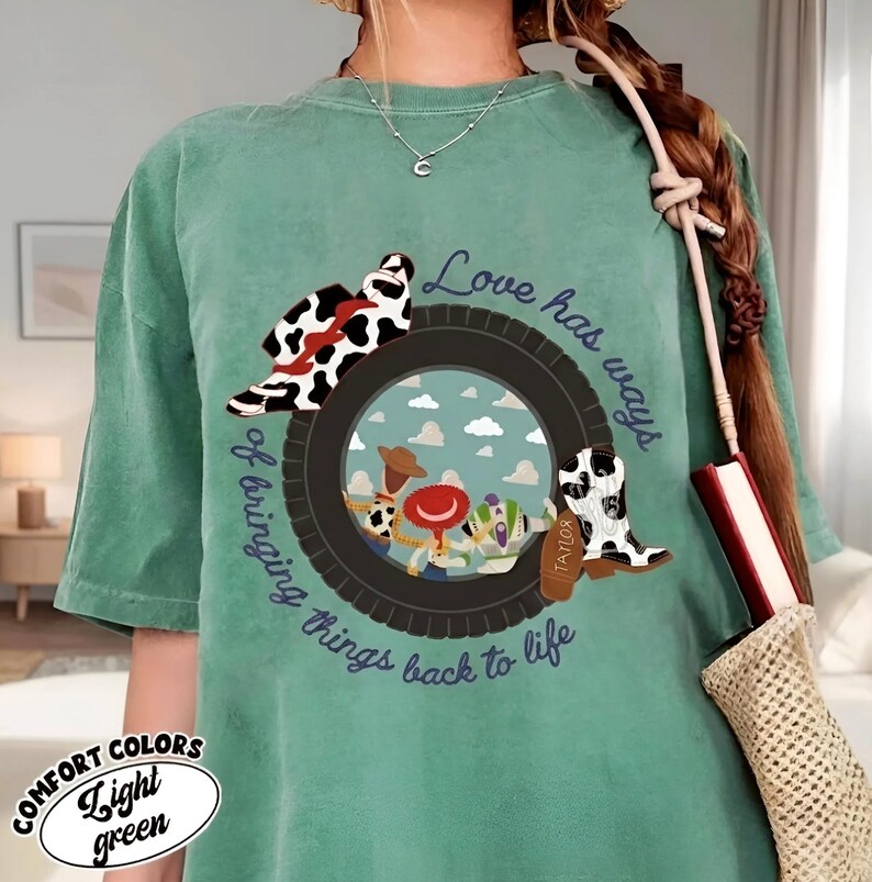 Puede incluir: Una camiseta verde claro con un gr&aacute;fico de un neum&aacute;tico negro que enmarca una escena de cielo con tres personajes de dibujos animados. Un sombrero y una bota con estampado de vaca est&aacute;n sobre el neum&aacute;tico. El texto dice, Love has ways of bringing things back to life.