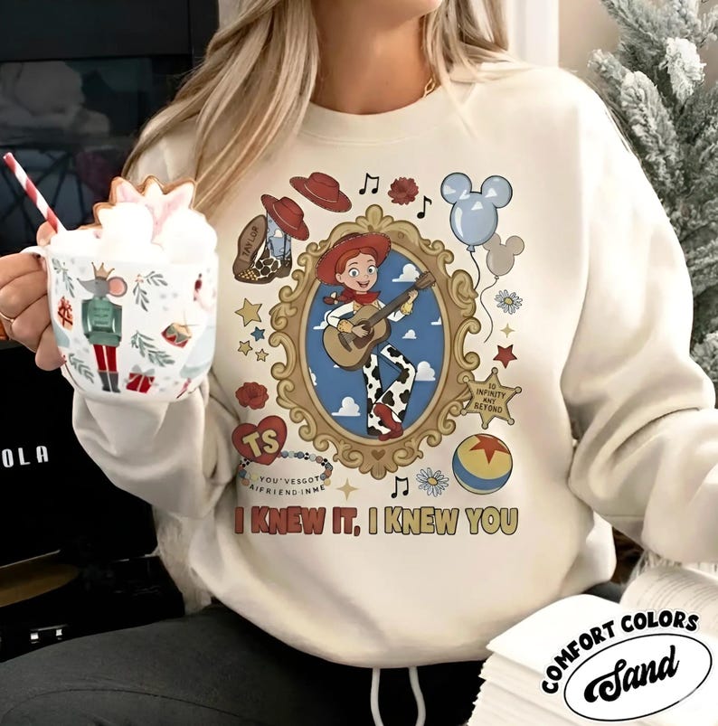 May include: A sand-colored crewneck sweatshirt featuring a central graphic of a cowgirl character playing a guitar inside an ornate gold frame. Surrounding elements include a red hat, a blue balloon, a star badge, and the text I KNEW IT, I KNEW YOU.