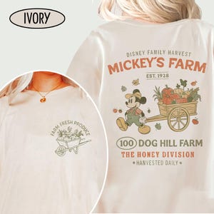 May include: Ivory-colored t-shirt featuring a graphic of Mickey Mouse with a cart of pumpkins, with the text "MICKEY'S FARM" and "EST. 1928." The front of the shirt has a wheelbarrow graphic with the words "FARM FRESH PRODUCE."