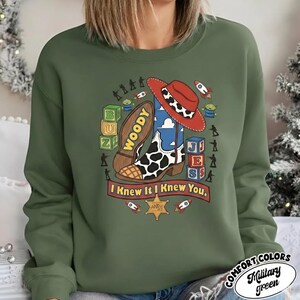 May include: A military green crewneck sweatshirt featuring a graphic print of a cowboy boot, a red hat, alphabet blocks, and small toy figures. The text "WOODY" is on the boot sole, and a banner below reads "I Knew It I Knew You" with a sheriff star.