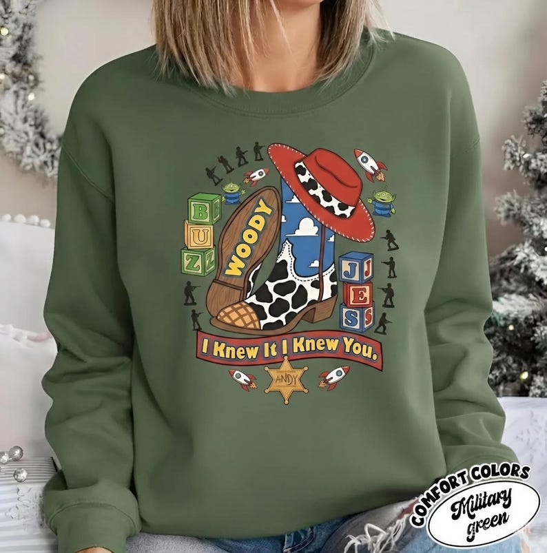 May include: A military green crewneck sweatshirt featuring a graphic print of a cowboy boot, a red hat, alphabet blocks, and small toy figures. The text "WOODY" is on the boot sole, and a banner below reads "I Knew It I Knew You" with a sheriff star.