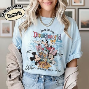 May include: Light blue Comfort Colors Chambray t-shirt featuring a Walt Disney World graphic with various cartoon characters. The design includes the text "Disneyworld", "est. 1971", and "Where dreams come true".
