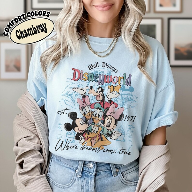 May include: Light blue Comfort Colors Chambray t-shirt featuring a Walt Disney World graphic with various cartoon characters. The design includes the text "Disneyworld", "est. 1971", and "Where dreams come true".