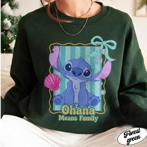 May include: Forest green sweatshirt featuring a blue cartoon character with large ears and the text "Ohana Means Family." The character is set against a striped background with a pink seashell and a blue bow.