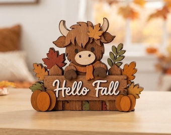 May include: A layered wooden decorative sign featuring a brown Highland cow with a scarf, positioned behind a fence. The sign displays the text Hello Fall in white script, accented with orange and red maple leaves and two orange pumpkins.