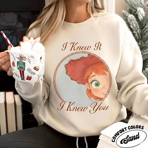 May include: A cream-colored crewneck sweatshirt featuring a circular graphic of a red-haired character with green eyes, framed by a rope border. The text "I Knew It" appears above and "I Knew You" below the graphic in a brown serif font.