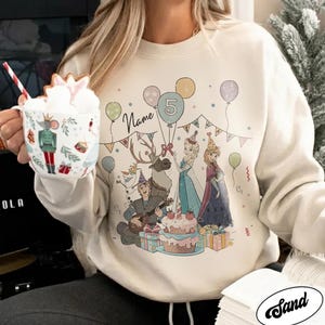 Puede incluir: Sudadera de color crema con un gr&aacute;fico colorido de personajes de dibujos animados, globos y una tarta de cumplea&ntilde;os. El dise&ntilde;o incluye el n&uacute;mero 5 y espacio para un nombre. Un dise&ntilde;o festivo para una celebraci&oacute;n de cumplea&ntilde;os.