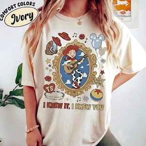 May include: An ivory t-shirt featuring a graphic print of a cowgirl character playing a guitar inside an ornate gold frame. The design includes a cowboy hat, a boot, a star badge with text, a ball, balloons, and the text I KNEW IT, I KNEW YOU.