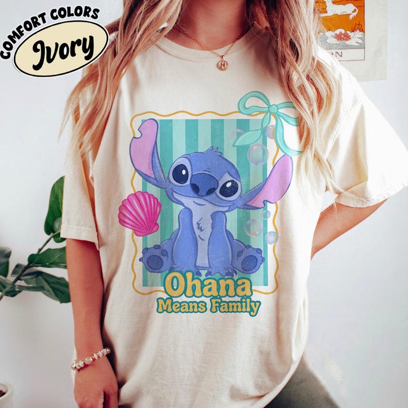 May include: Ivory-colored t-shirt featuring a blue cartoon character with large ears and the text "Ohana Means Family." The design includes a pink seashell and a teal bow. The words "Comfort Colors" and "Ivory" are printed on the top left.