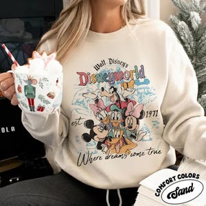 May include: Cream-colored sweatshirt featuring a colorful graphic of Disney characters, including Mickey Mouse, Minnie Mouse, Donald Duck, and Goofy. The design includes the words "Walt Disney's Disneyworld" and "Where dreams come true."