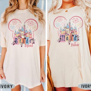 Può includere: Due magliette color avorio con una grafica di un castello rosa e blu con fuochi d'artificio e personaggi di Topolino. Il testo sulle magliette recita "Bride Squad" e "Maid of Honor".