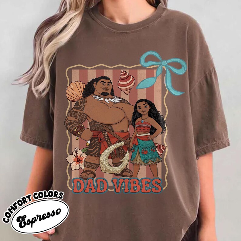 May include: Espresso-colored t-shirt featuring a graphic of two animated characters with the text "DAD VIBES." The design includes a light blue bow, a flower, and seashell accents. The shirt is part of the Comfort Colors collection.