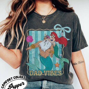 May include: Dark gray t-shirt featuring a cartoon illustration of King Triton and Ariel from The Little Mermaid, with the text "DAD-VIBES". The shirt also has the Comfort Colors and Pepper logos.