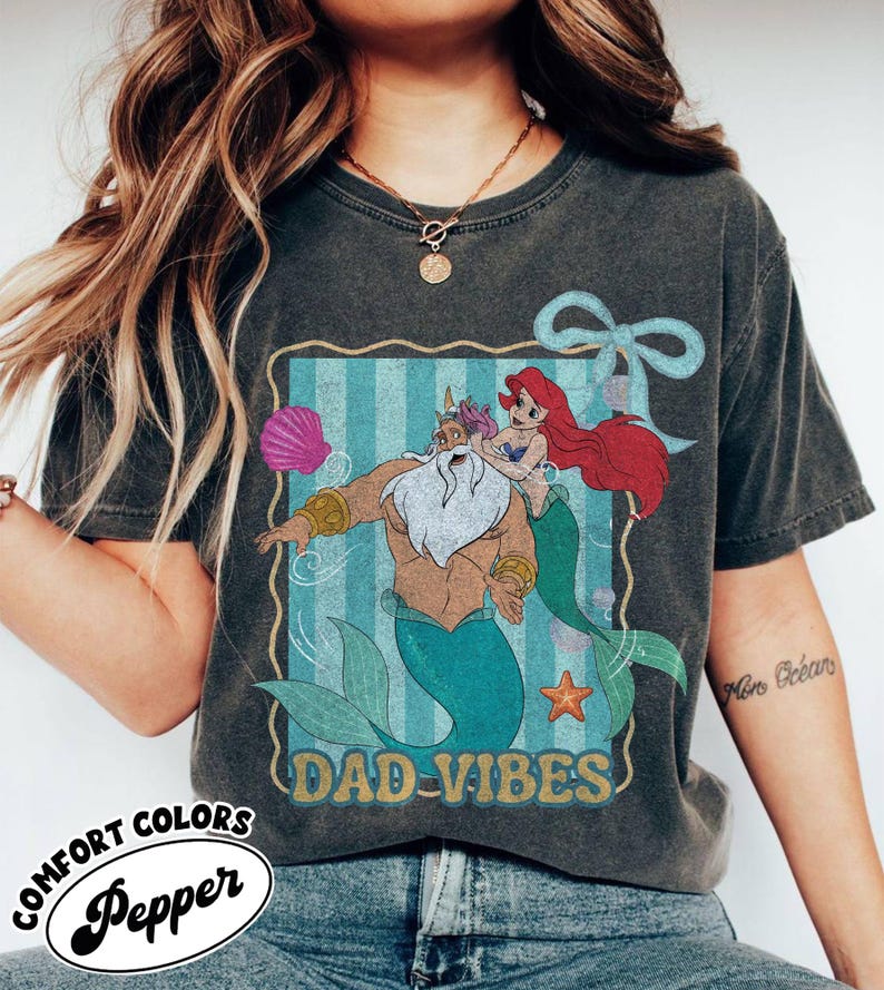 May include: Dark gray t-shirt featuring a cartoon illustration of King Triton and Ariel from The Little Mermaid, with the text "DAD-VIBES". The shirt also has the Comfort Colors and Pepper logos.