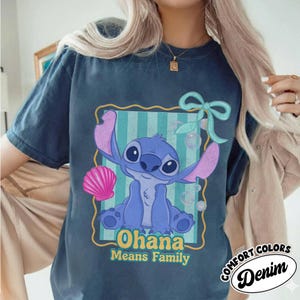 May include: A navy blue t-shirt featuring a cartoon illustration of Stitch from Lilo & Stitch. The design includes the words "Ohana Means Family" and a seashell. The shirt has a distressed look and the words "Comfort Colors Denim" are printed on the bottom right.