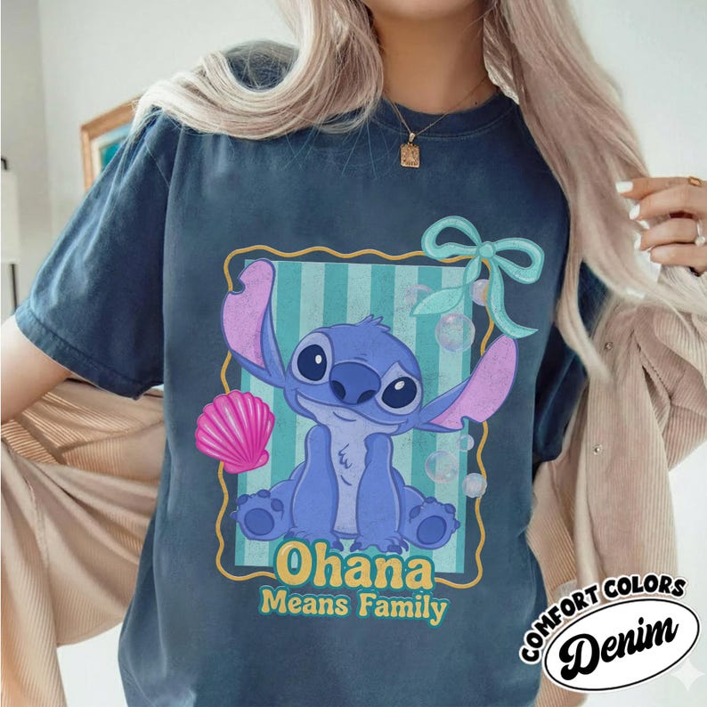 May include: A navy blue t-shirt featuring a cartoon illustration of Stitch from Lilo & Stitch. The design includes the words "Ohana Means Family" and a seashell. The shirt has a distressed look and the words "Comfort Colors Denim" are printed on the bottom right.