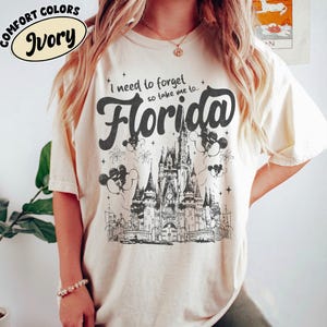 May include: Ivory t-shirt featuring the text "I need to forget so take me to... Florida" in black script. The design includes a detailed illustration of a castle, fireworks, and Mickey Mouse balloons. The shirt is labeled "Comfort Colors".