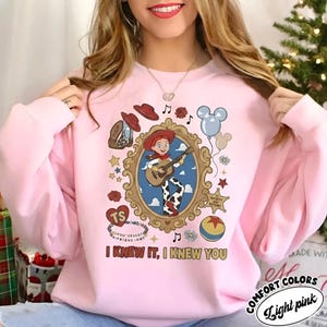 May include: A light pink sweatshirt featuring a graphic print of a cartoon cowgirl playing a guitar inside an ornate gold frame. Surrounding the frame are icons including a cowboy hat, a boot, a balloon, a ball, and the text I KNEW IT, I KNEW YOU.