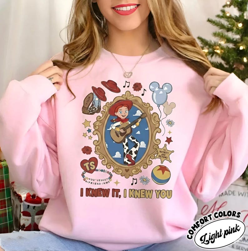 May include: A light pink sweatshirt featuring a graphic print of a cartoon cowgirl playing a guitar inside an ornate gold frame. Surrounding the frame are icons including a cowboy hat, a boot, a balloon, a ball, and the text I KNEW IT, I KNEW YOU.
