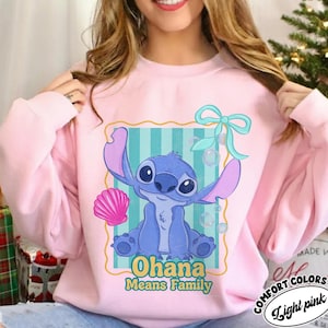 May include: Light pink sweatshirt featuring a blue cartoon character with large ears and the text "Ohana Means Family." The design includes a pink seashell, bubbles, and a teal bow. The sweatshirt is labeled "Comfort Colors" and "Light Pink."