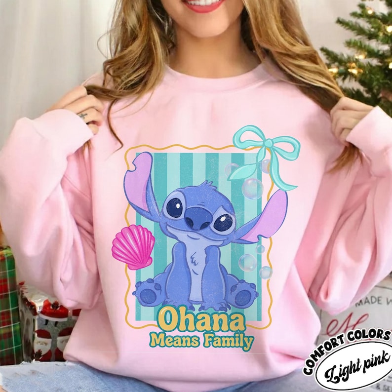 May include: Light pink sweatshirt featuring a blue cartoon character with large ears and the text "Ohana Means Family." The design includes a pink seashell, bubbles, and a teal bow. The sweatshirt is labeled "Comfort Colors" and "Light Pink."