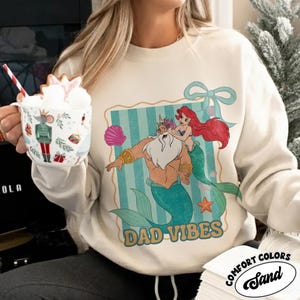 May include: Cream-colored sweatshirt featuring a cartoon illustration of King Triton and Ariel from The Little Mermaid, with the text "DAD VIBES". The design includes a seashell and a bow. The sweatshirt is from Comfort Colors.