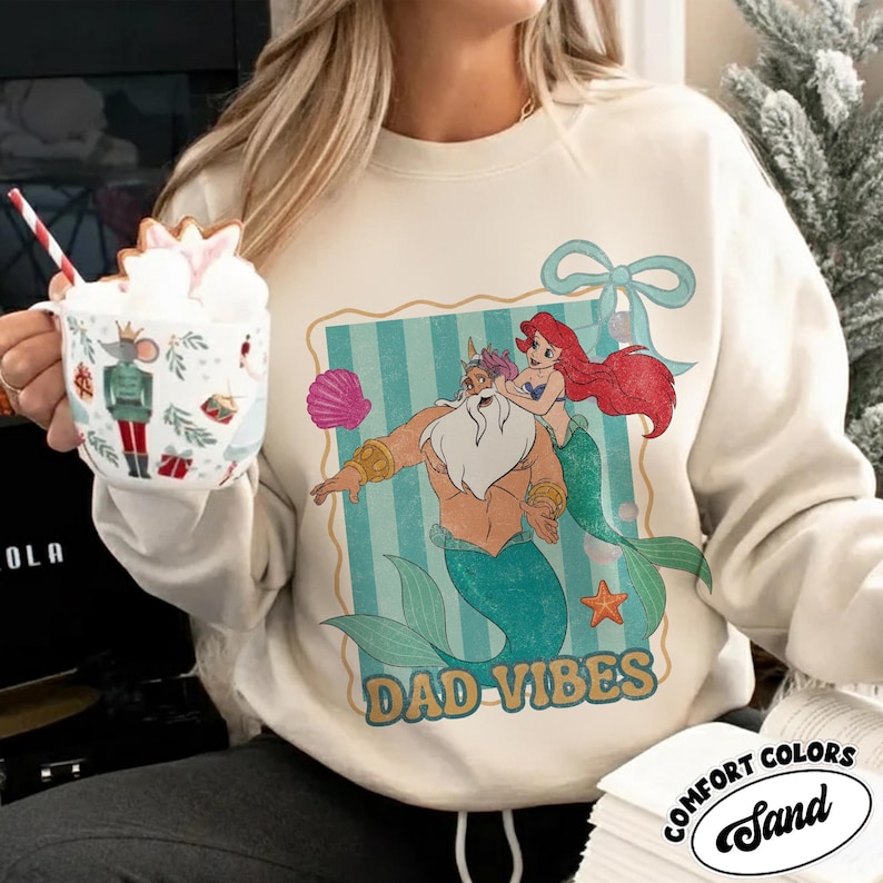 May include: Cream-colored sweatshirt featuring a cartoon illustration of King Triton and Ariel from The Little Mermaid, with the text "DAD VIBES". The design includes a seashell and a bow. The sweatshirt is from Comfort Colors.
