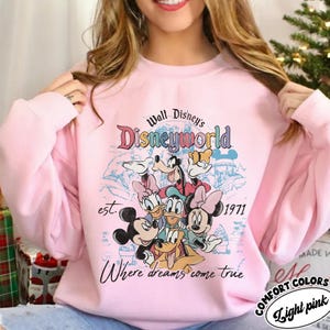 May include: Light pink sweatshirt featuring a Disneyworld graphic with Mickey Mouse, Minnie Mouse, Donald Duck, Daisy Duck, Goofy, and Pluto. The design includes the text "Walt Disney's Disneyworld", "est. 1971", and "Where dreams come true".